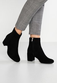 Černé semišové kotníkové boty se čtveratou špičkou a blokovým podpatkem. Mají boční zip pro snadné obouvání. Kombinované se šedými kostkovanými tapered kalhotami.