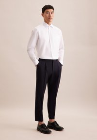 Chemise blanche à manches longues avec un col boutonné et une poche poitrine, associée à un pantalon noir marine ajusté et des chaussures à lacets noires.