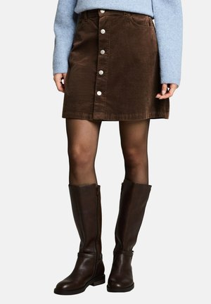 Femme portant une jupe boutonnée en velours côtelé marron, des collants noirs transparents, des bottes en cuir marron foncé montantes jusqu'aux genoux et un pull bleu clair.