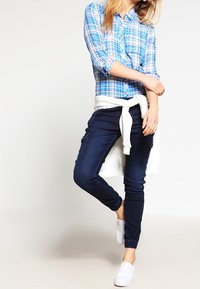 Chemise à carreaux bleu et blanc avec manches retroussées, associée à un pantalon cargo slim bleu foncé et des chaussures blanches sans lacets.