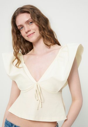 Jeune femme aux cheveux bruns ondulés portant un haut sans manches crème avec de larges volants aux épaules et un lien devant, souriant légèrement.