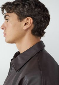 Bruine leren jas met een kraag, zichtbare stiksels en een gladde textuur, getoond op een model van opzij.