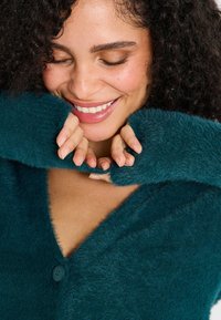 Cárdigan esponjoso en color verde azulado con un profundo escote en V, que cuenta con un cierre de botones. La textura suave es evidente, con mangas largas parcialmente arremangadas.