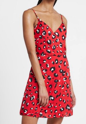 Femme portant une robe rouge sans manches avec un imprimé léopard noir et blanc et des fines bretelles, debout devant un fond uni.