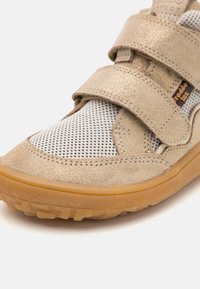 Pantofii din suede auriu deschis cu partea superioară din plasă, două benzi velcro și talpă din cauciuc. Remarcabili pentru finisajul texturat și designul sport.