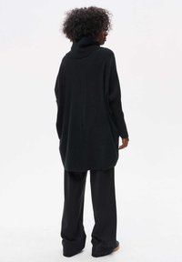 Oversized zwart gebreid sweatervan met hoge kraag, lange mouwen en gestructureerde stof. Gecombineerd met losse zwarte broeken, gezien vanaf de achterkant.