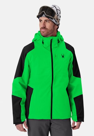 Homme portant une veste de ski verte et noire, un pantalon gris, un bonnet blanc à motifs et des lunettes de ski regardant sur le côté sur un fond blanc.