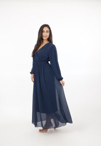 Vrouw in een navyblauwe doorschijnende jurk met lange mouwen poseert met één hand die de rok vasthoudt, draagt transparante hoge sandalen, tegen een witte achtergrond.