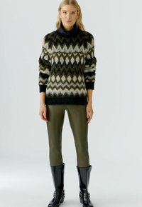 Multi-kleurige wollen trui met een zwarte col, voorzien van een zigzagpatroon in groen en crème. Gepaarde met olijfgroene leggings en zwarte laarzen.