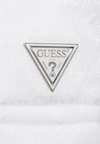 Weißes, strukturiertes Stofflogo-Patch mit einem silbernen dreieckigen Design, versehen mit dem Text "GUESS" und "WASHED JEANS" sowie einem Fragezeichen-Detail.