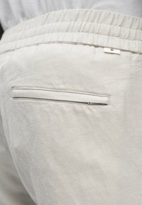 Lichtgrijze broek met een elastische tailleband, een enkele achterzak met rits, en een zachte, gladde stoftextuur.