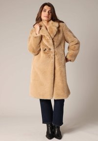 Abrigo de piel sintética beige con un amplio cuello, cierre de doble botón y un ajuste relajado. Lleva puesto sobre jeans oscuros y botas de tobillo.