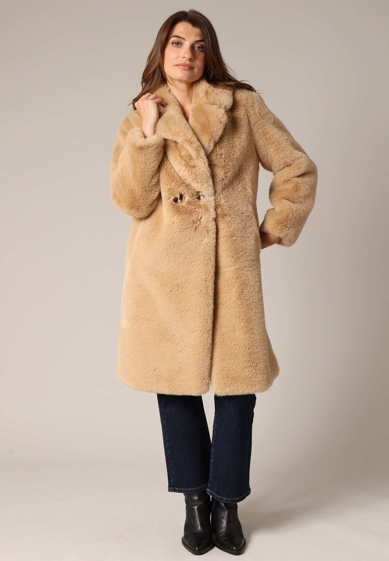 Abrigo de piel sintética beige con un amplio cuello, cierre de doble botón y un ajuste relajado. Lleva puesto sobre jeans oscuros y botas de tobillo.