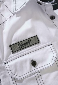 Valge kangast särk, millel on kontrastsed mustad õmblused, tasku tumeda nööbiga ja tikandiga silt "Brandit" kalligraafias.