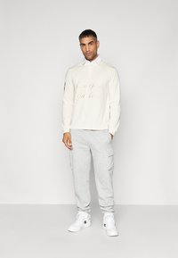 White pullover with "New York Yankees" text, gray cargo pants, and white sneakers. Features a collared neckline and long sleeves.