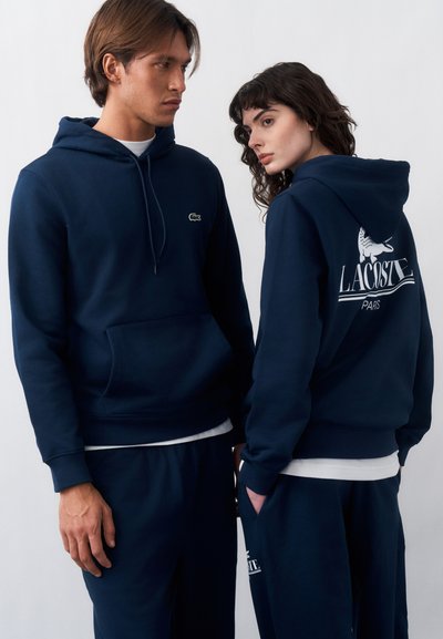 Мъж и жена, облечени с еднакви тъмносини суичъри и панталони Lacoste, жената показва голямо бяло лого на Lacoste на гърба на суичъра.