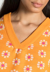 V-ringad tröja i ljus orange stickat tyg med vita och rosa blommönster. Ribbad krage som tillför textur till designen.