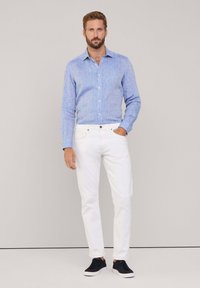 Lichtblauw overhemd met een subtiele textuur, wit slim-fit jeans en zwarte sneakers, staand tegen een neutrale achtergrond.
