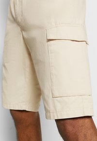 Raffinati pantaloni cargo beige realizzati in tessuto testurizzato, con tasche laterali e una linea dritta, con orlo che raggiunge la metà della coscia.