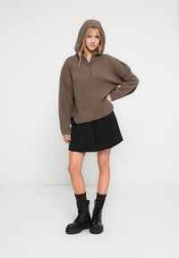 Γυναίκα που φοράει καφέ φλις με κουκούλα, μαύρη πλισέ φούστα, σκούρες κάλτσες και μαύρα μποτάκια στρατιωτικού τύπου με κορδόνια, στέκεται με το ένα χέρι στη μέση.