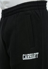 Οι μαύρες φόρμες διαθέτουν ριμπ ελαστική μέση, πλαϊνές τσέπες και λευκό λογότυπο "CARHARTT". Το ύφασμα φαίνεται μαλακό και με υφή.