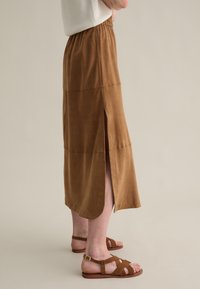 Jupe midi en suedine marron avec fentes latérales, taille élastique et texture lisse. Associée avec des sandales à brides marron.