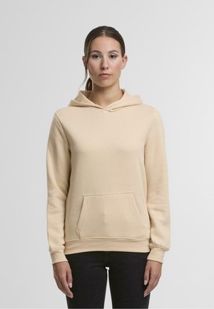 Un hoodie beige en tissu doux, avec une poche avant, une capuche réglable et des poignets côtelés. Porté avec un jean foncé.