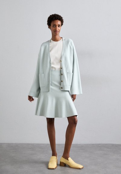 Lys mintgrønn cardigan med to frontlommer over et matchende figursydd skjørt. Båret med teksturerte gule slip-on sko og en hvit topp.