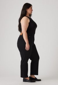 Un débardeur noir côtelé associé à un jean noir taille haute. Le jean a une légère évasement, cinq poches et une étiquette en cuir de marque à l'arrière.