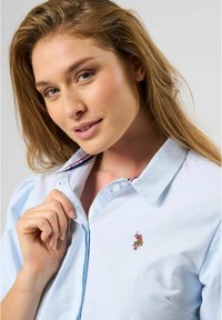 Light blue button-up shirt with a collar, featuring contrasting striped interior, and an embroidered logo on the chest. Soft fabric texture.