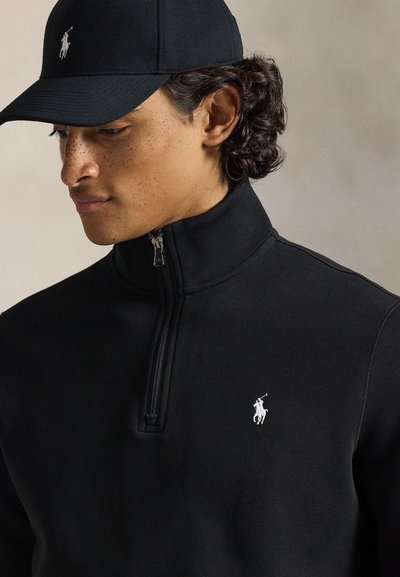 Svart jakke med glidelås, høy krage og hvit polo-logo. Modellen har også på seg en matchende svart caps. Glatt stoff og tettsittende design.