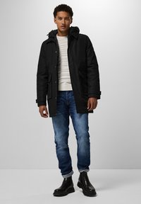 Černá kapuce parka se zipem a patkami, s dvěma předními kapsami, nošená přes světle pruhovaný svetr, spárovaná s modrými džínami a černými botami.