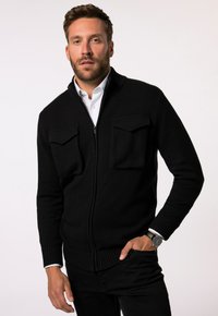Schwarzer gestrickter Reißverschluss-Pullover mit zwei Vordertaschen, gerippten Bündchen und Saum, getragen über einem weißen Hemd mit Kragen. Flache Textur, lässiges Design.