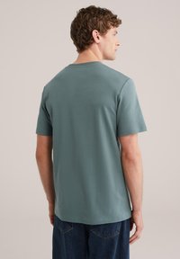 Man met krullend bruin haar, gekleed in een gedempte groene T-shirt met korte mouwen en donkerblauwe spijkerbroek, staand met de rug naar de camera tegen een beige achtergrond.