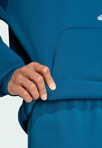 Tealfärgad sweatshirt med ribbad fåll och ficka. Hand som greppar fållen och visar den mjuka tygtexturen och den sömlösa designen.