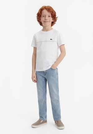 Băiat cu păr roșcat, stând cu o mână în buzunar, purtând un tricou alb cu logo, blugi bleu deschis și pantofi slip-on bej.