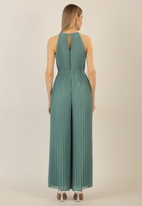 Pleated jumpsuit in muted green with a high neck and keyhole back, featuring an elastic waist and wide-leg design. Silver heeled shoes visible.