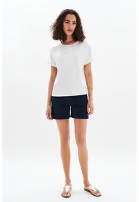 Top blanco de manga corta con puños enrollados, combinado con pantalones cortos azul marino con bolsillos frontales y un ajuste relajado; el modelo lleva sandalias metálicas.
