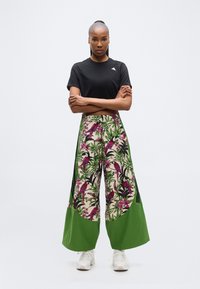 Mujer de pie con los brazos cruzados, lleva una camiseta negra corta y pantalones de pierna ancha con estampado tropical de aves verdes y rosas, zapatillas blancas.
