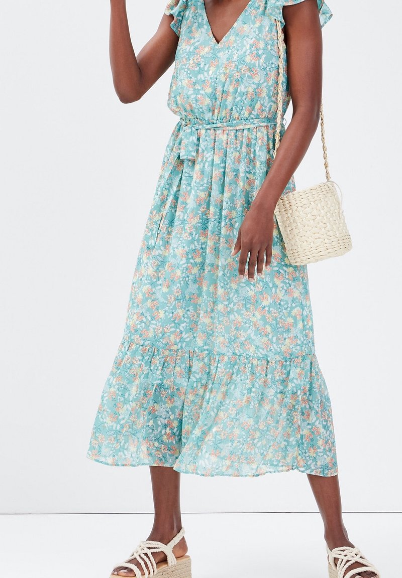 Femme portant une robe midi bleu clair à motifs floraux avec des manches à volants, une ceinture à nouer à la taille, des sandales en corde blanches, et portant un petit sac bandoulière tressé crème.