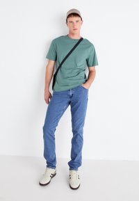Camiseta de algodón verde combinada con jeans azules. El modelo lleva una gorra beige y zapatillas ligeras con acentos oscuros. Conjunto simple y casual.