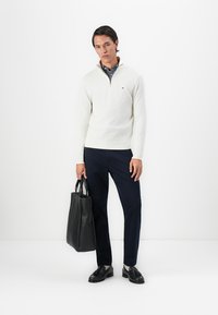 Mann iført hvit kvart-draget genser, marineblå bukser, svarte frakkeskinnslipper, holder en stor svart skinn tote-bag, står mot en hvit bakgrunn.