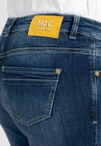 Jean en denim bleu foncé avec une étiquette en cuir jaune doré portant la mention « MAC » à la ceinture. Dispose de poches arrière avec des coutures apparentes.