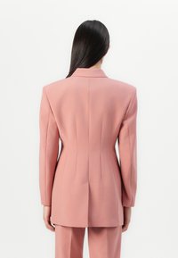 Růžový přizpůsobený blazer se strukturovaným střihem, ostrými klopami a detailem švu na zadní straně; hladká tkanina s matným povrchem.