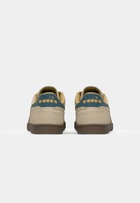 Tenis de camurça bege com uma sola de borracha castanha escura, apresentando um detalhe em teal no calcanhar e uma marcação dourada na parte de trás.