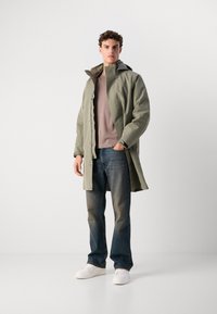 Abrigo impermeable verde claro con capucha, superpuesto a un suéter rosa apagado, combinado con jeans azul oscuro y zapatillas blancas.