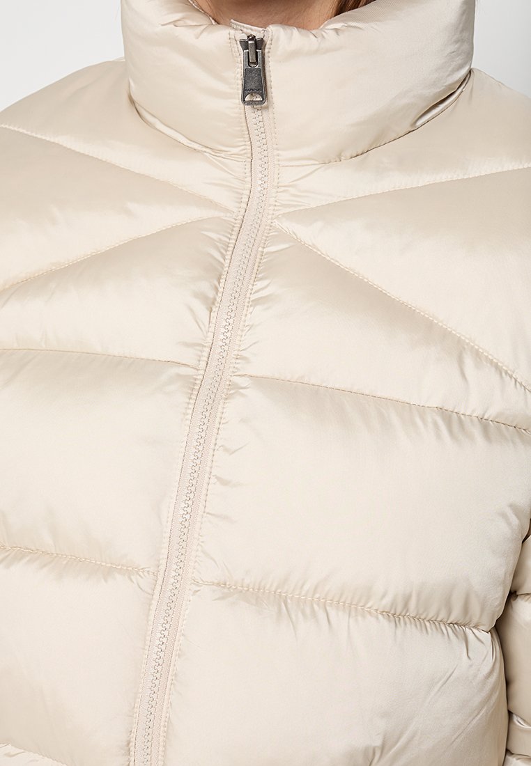Zbliżenie na kremową, pikowaną kurtkę puchową z wysokim kołnierzem i centralnym zamkiem, noszoną przez osobę.