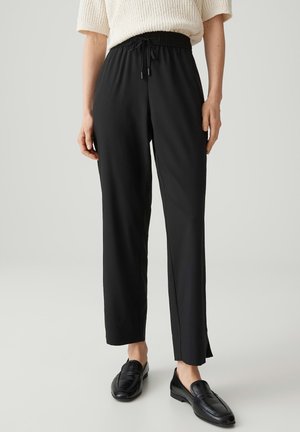 Frau trägt schwarze, hoch taillierte Hose mit Kordelzug, cremefarbenes Kurzarm-Stricktop und schwarze Loafers, steht auf einem hellgrauen Boden.