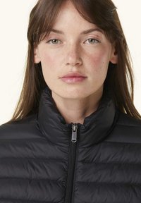 Giubbotto imbottito nero con colletto alto, dotato di zip frontale e texture trapuntata, indossato da una persona con lunghi capelli castani.