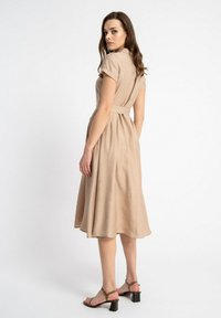 Beżowa sukienka z kołnierzykiem i krótkimi rękawami, zebrana w talii, z luźną spódnicą i subtelną teksturą; noszona z sandałami na słupku z paskami.